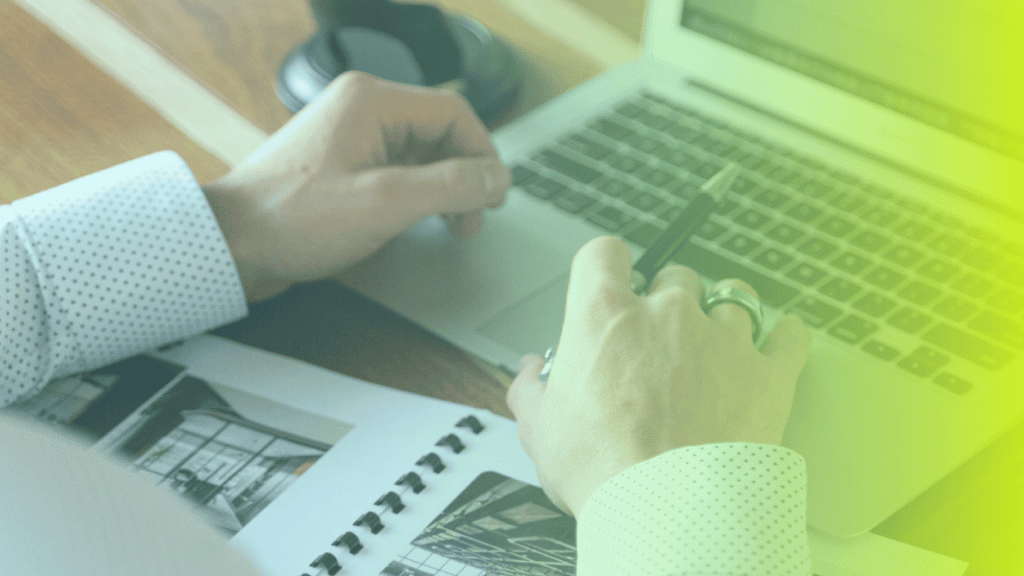 A person wearing a white shirt works on a laptop with an open notebook containing architectural sketches on a wooden desk—an inspiring scene that reflects the importance of corporates wellness in creative workspaces.