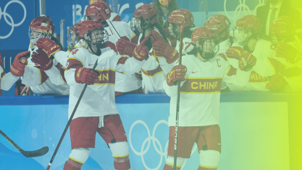 Chinese womens ice hockey players in white and red uniforms celebrate on the ice in front of their team bench during an Olympic event, showcasing teamwork and inspiration that can benefit corporates wellness programs.