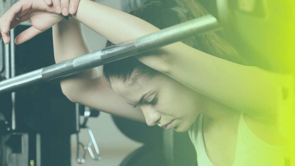 A woman rests her arms and head on a barbell in a gym, appearing tired or contemplative—a moment highlighting the importance of corporates wellness. The image features a greenish-yellow gradient overlay.