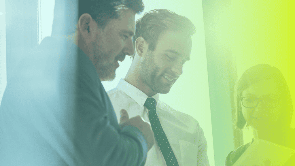 Three people in business attire stand together indoors, looking at a tablet and smiling, with soft blue and yellow light overlaying the image—capturing a moment of corporates wellness in the workplace.