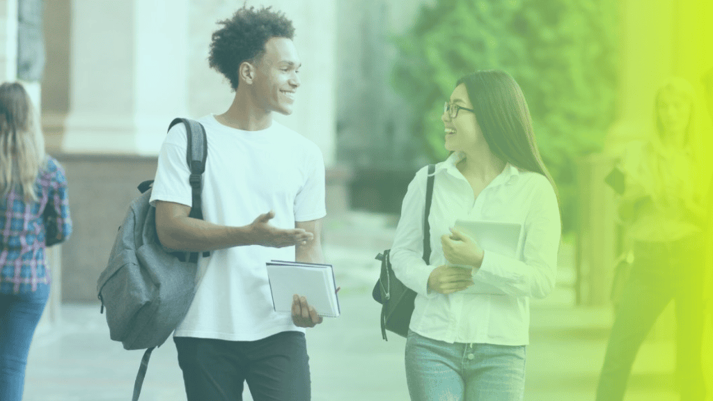 Two students walk together outdoors, smiling and talking about corporate wellness. One holds notebooks, the other holds a tablet. Trees and other people are in the background.