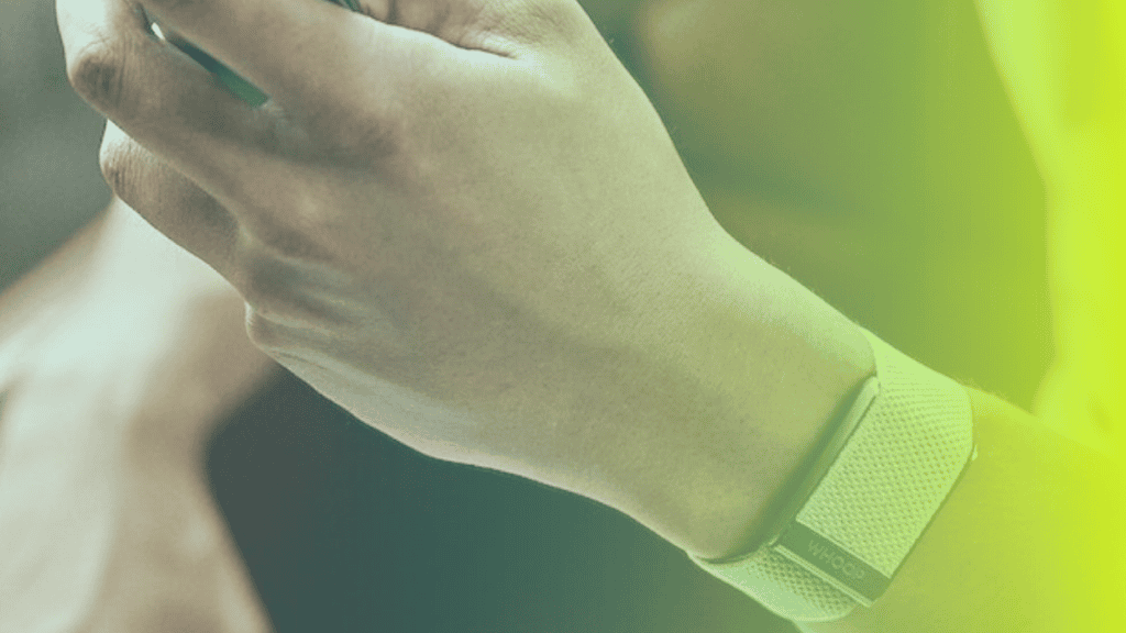 A close-up of a person’s hand holding a device, wearing a textured wristband on their left wrist. The image, with its green and yellow gradient overlay, reflects a modern corporates wellness approach.