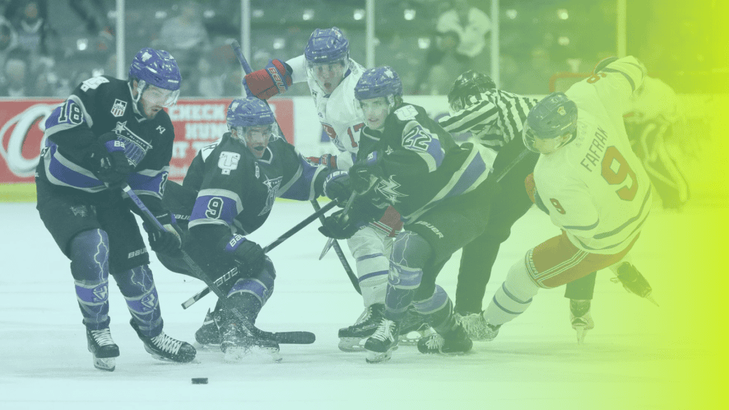 Five ice hockey players and a referee engage near the puck during a game, with stands and corporates wellness advertisements visible in the background.