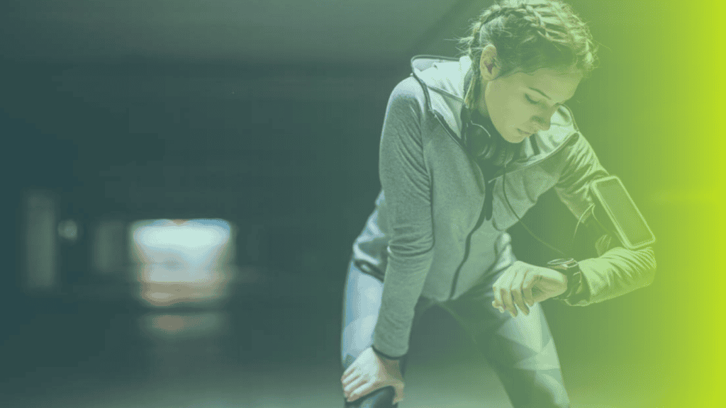 A woman in athletic wear checks her smartwatch while resting, with headphones around her neck and a smartphone armband, reflecting modern corporates wellness habits in a dimly lit setting.