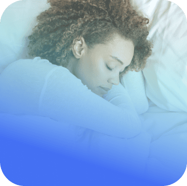 A woman with curly hair sleeps on her side in a bed with white bedding, wearing a light-colored long sleeve top from Coya.