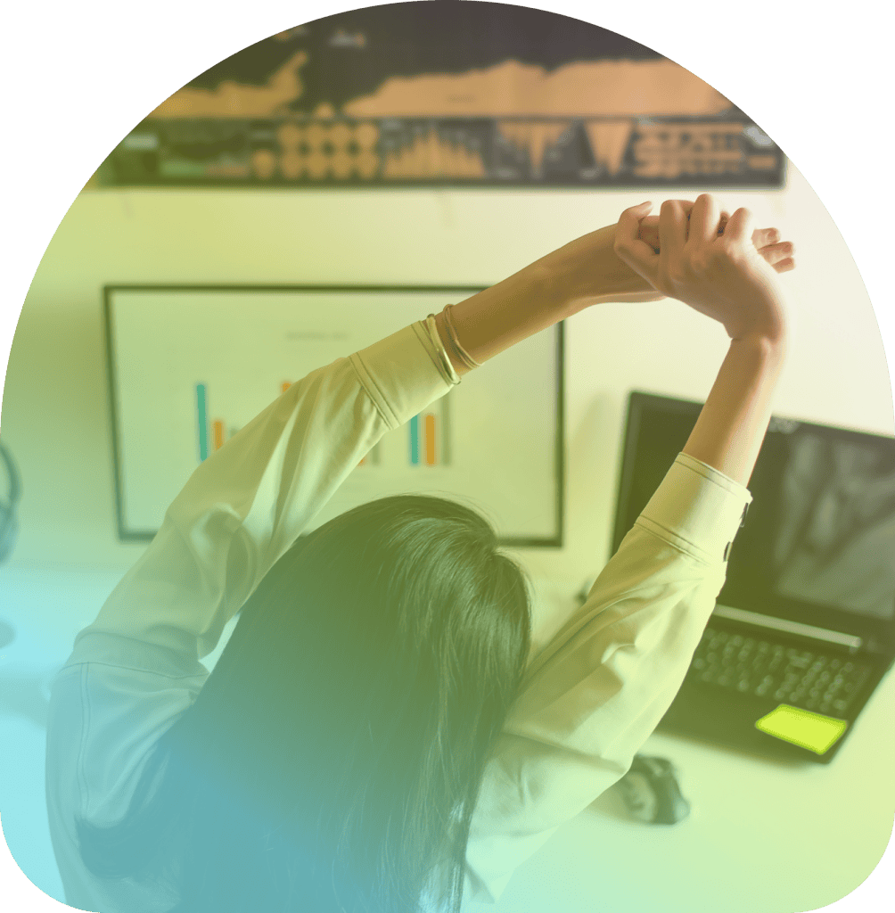 Person seated at a desk stretching arms overhead, with a laptop, monitor displaying graphs, and a wall chart in the background—an ideal setup for using Coya for companies to streamline data analysis and workflow.