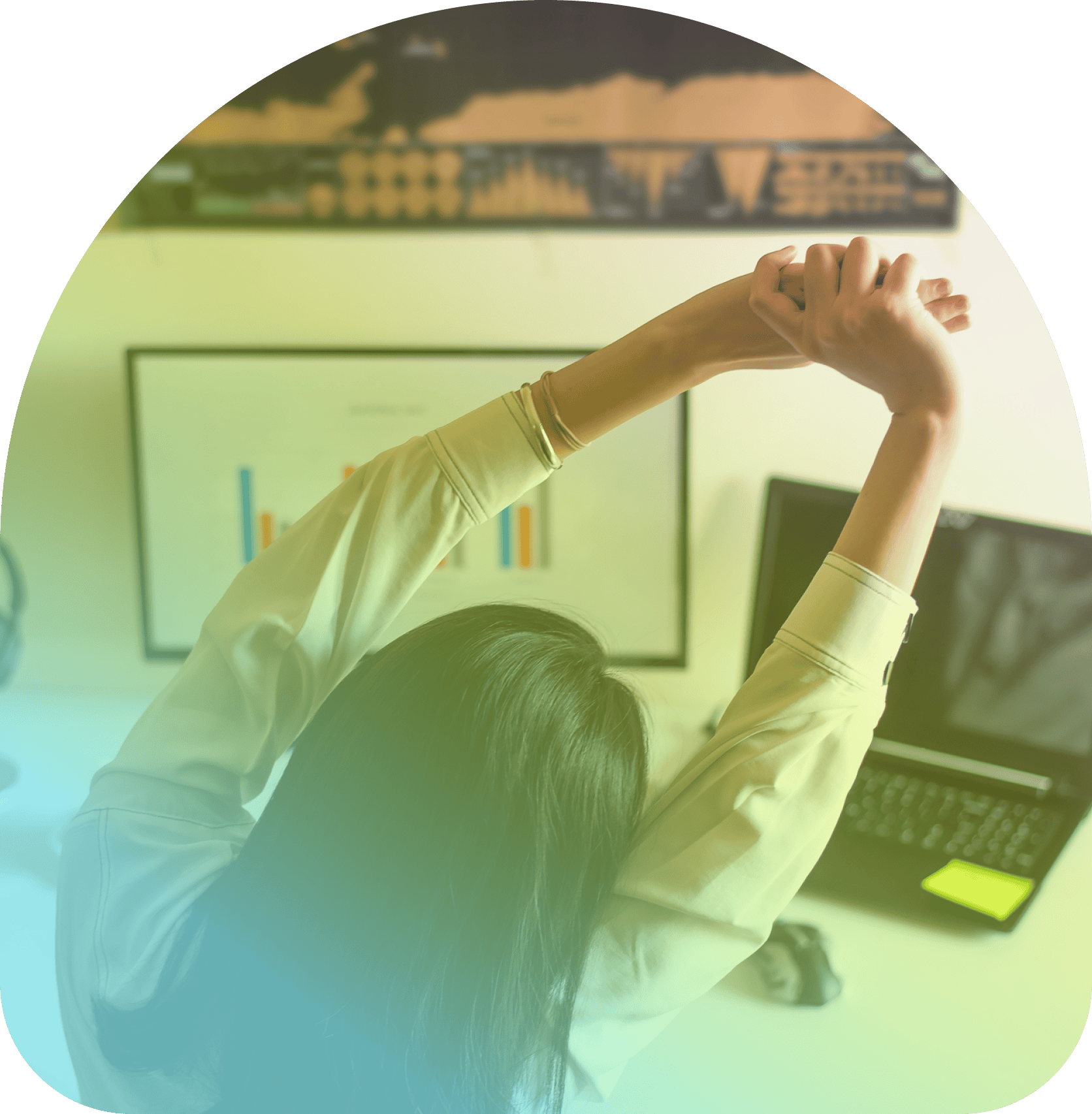 Person seated at a desk stretching arms overhead, with a laptop, monitor displaying graphs, and a wall chart in the background—an ideal setup for using Coya for companies to streamline data analysis and workflow.