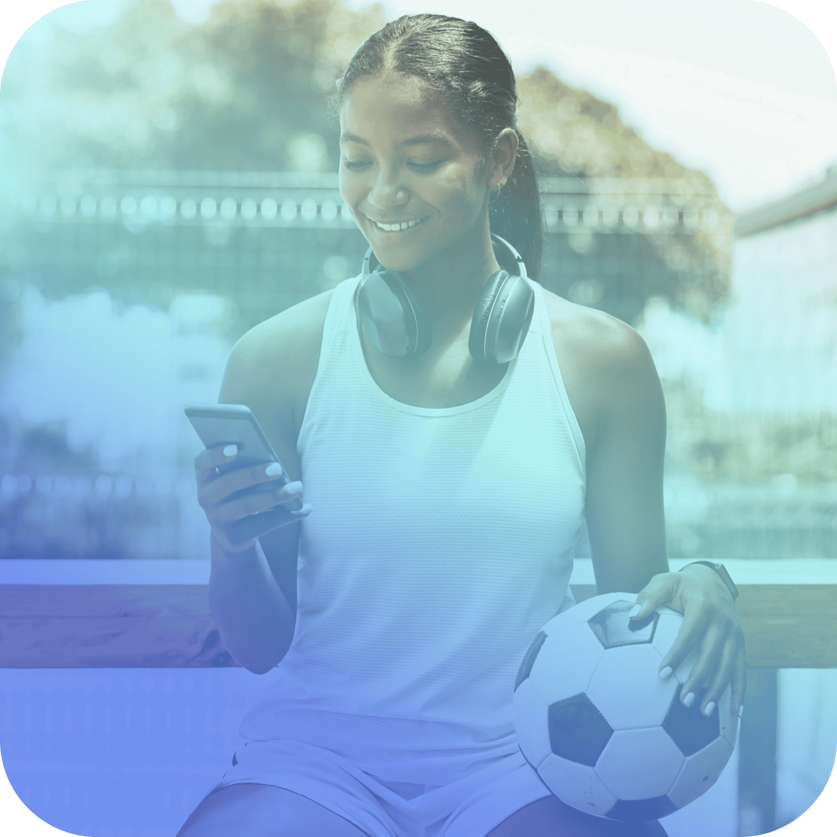 A person in sportswear with headphones around their neck sits outdoors, holding a soccer ball in one hand and checking coya for student athletes on their smartphone.