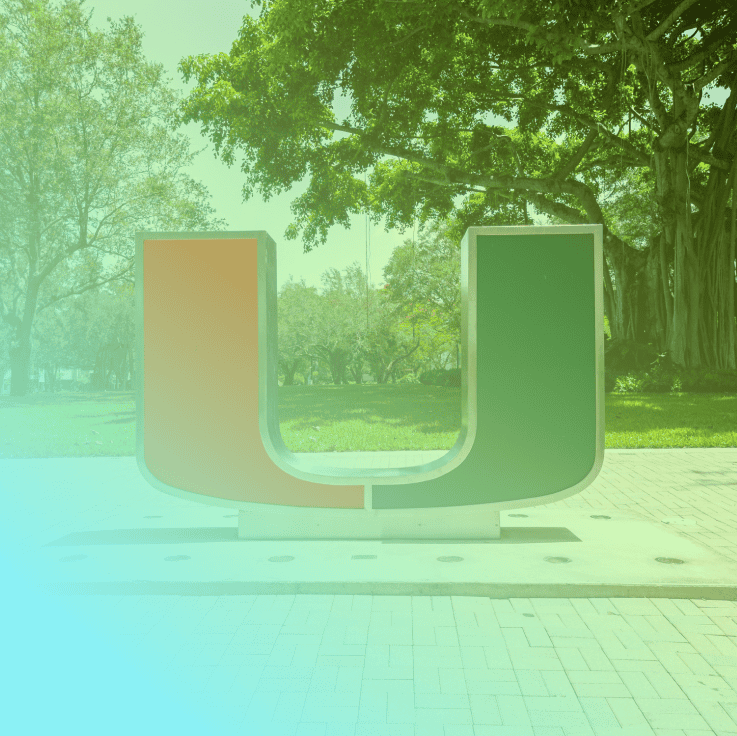 A large orange and green Coya U sculpture stands outdoors on a paved area, with trees and grass visible in the background.