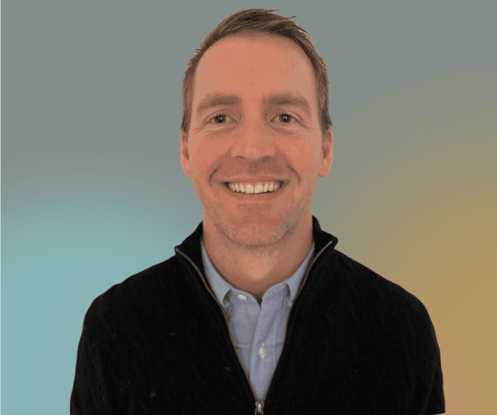 A man with short brown hair, wearing a light blue shirt and black zip-up sweater, smiles in front of a gradient blue and yellow background, photographed by Dayna McCutchin.