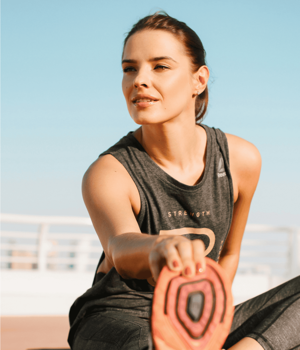 Woman wearing athletic clothes stretches her leg outdoors on a sunny day, holding her foot with one hand and looking into the distance—an active moment that captures the Coya for executives lifestyle.