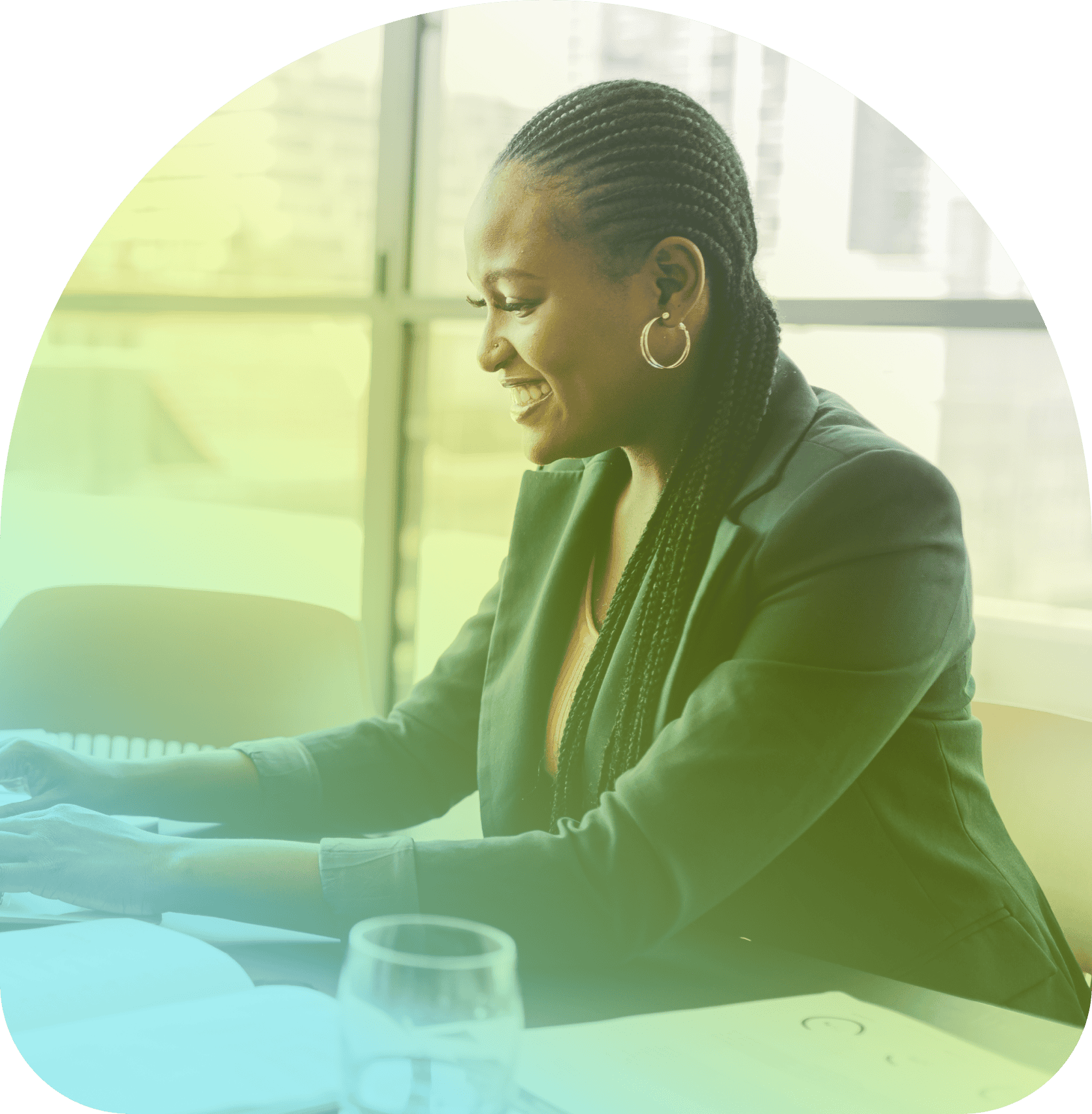 A woman in business attire sits at a desk, smiling while working on her Coya laptop. Papers and a glass of water are on the desk, with large windows in the background.