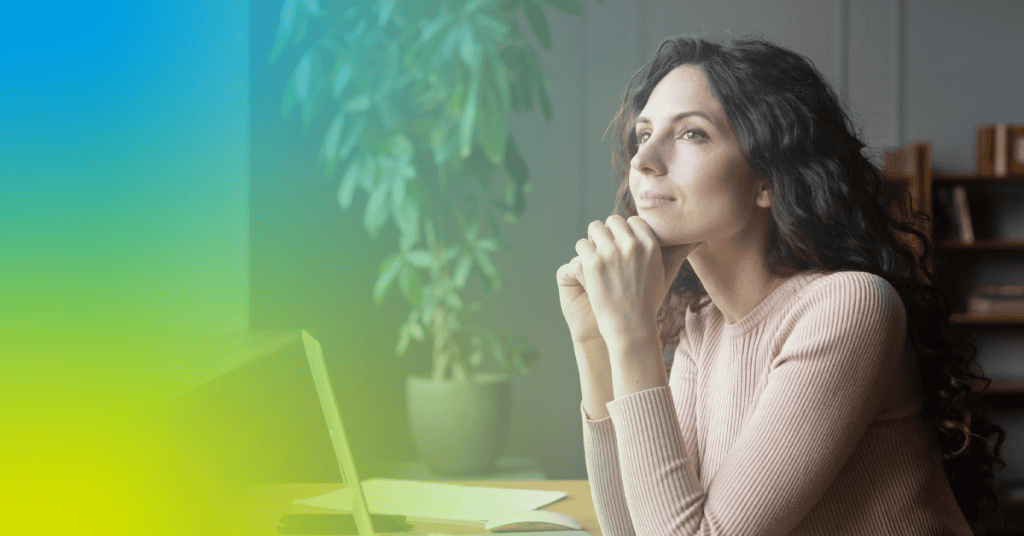 A woman with long, dark hair sits at a desk with a laptop, looking thoughtfully out a window, perhaps reflecting on stress. A potted plant and shelves are visible in the background.