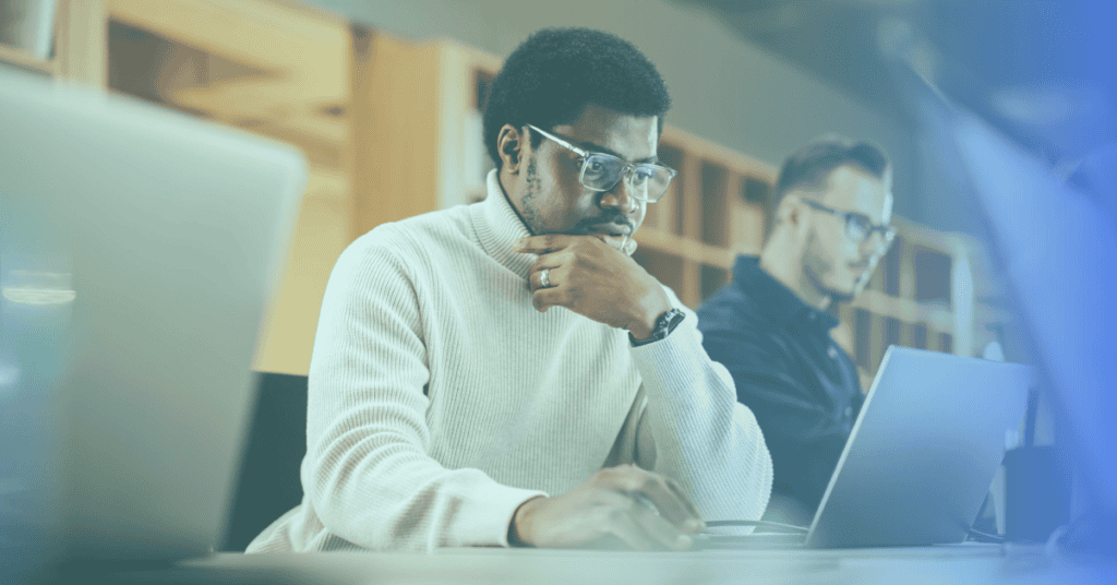 Two people are sitting at desks working on laptops in an office environment, demonstrating discipline as they remain focused on their screens.