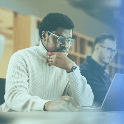 Two men sit at desks working on laptops in an office setting; one in the foreground, wearing glasses and a light sweater, displays discipline as he focuses intently on his screen.