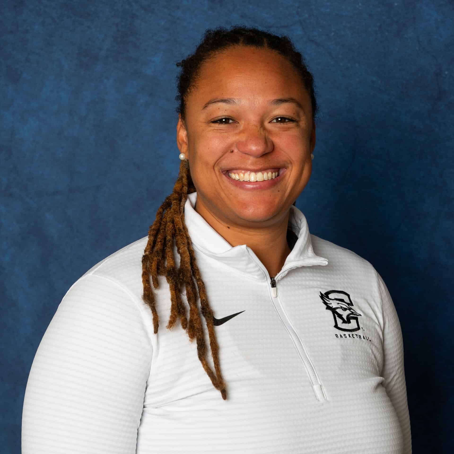 A person with long dreadlocks smiles at the camera, wearing a white zip-up athletic jacket with a basketball logo, against a blue backdrop, capturing Jordann Reeses transformative journey.