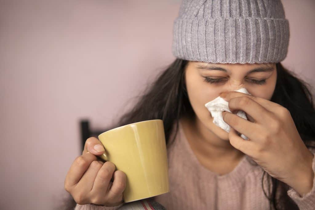 A person wearing a knit hat holds a yellow mug in one hand and uses a tissue to blow their nose, focusing on ways to strengthen their immune system.