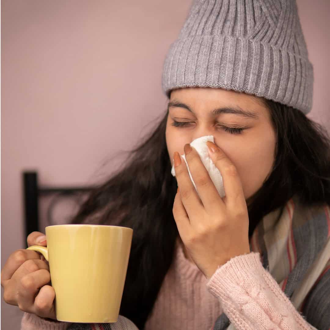 A person wearing a knit hat and scarf holds a yellow mug and covers their nose with a tissue, appearing to sneeze—suggesting the importance of keeping your immune system strong during cold weather.