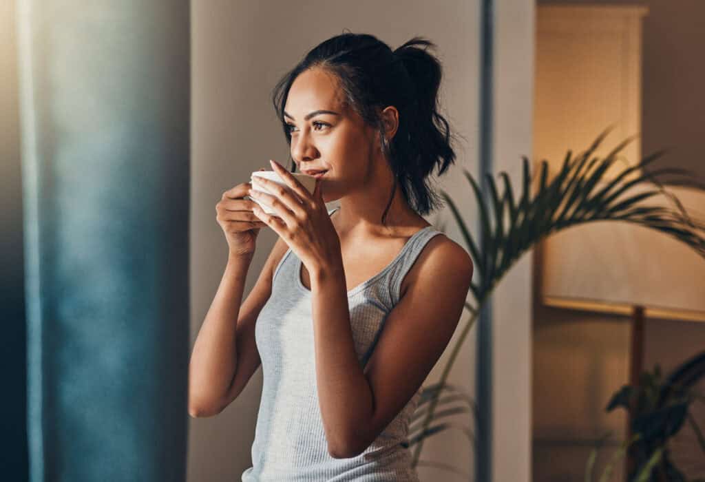 A young Hispanic woman enjoying a warm cup of coffee for breakfast. One mixed race female drinking tea while looking at the view from a window in her apartment