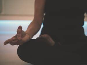 Person sitting cross-legged on the floor, wearing dark clothing, with one hand resting on their knee in a meditation gesture, embodying the purpose of meditation—finding inner calm and clarity.