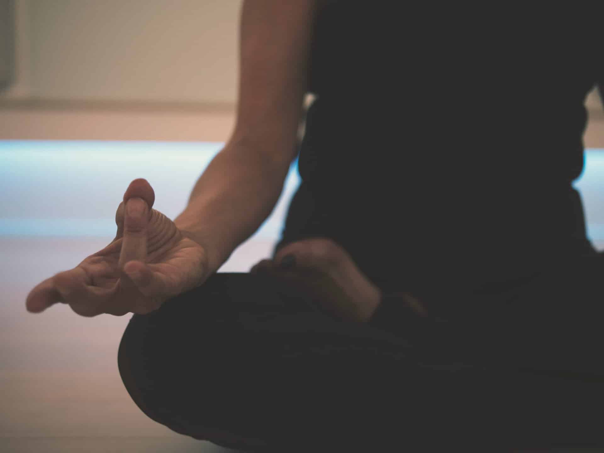 Person sitting cross-legged on the floor, wearing dark clothing, with one hand resting on their knee in a meditation gesture, embodying the purpose of meditation—finding inner calm and clarity.
